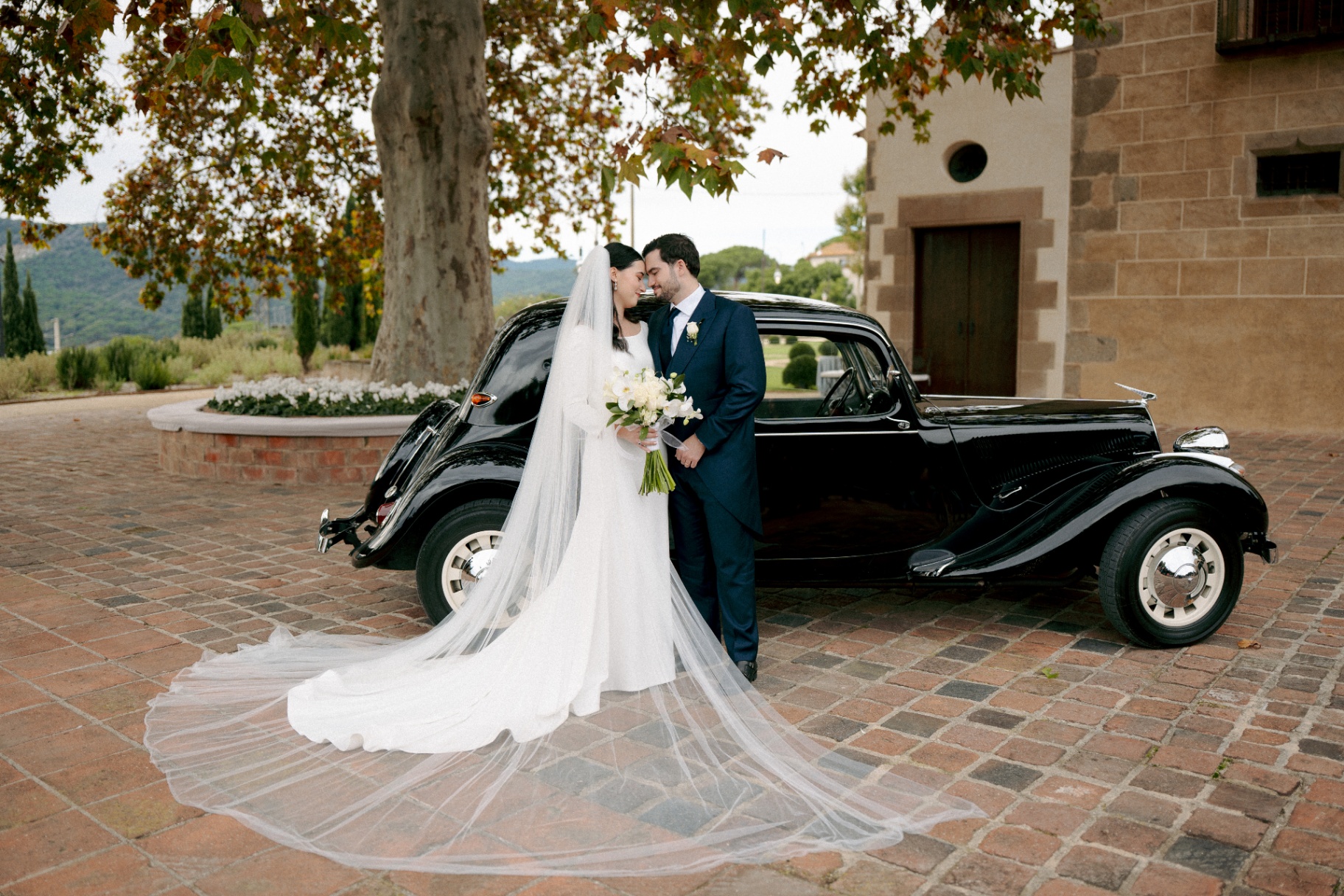 Helen y Pedro junto a un coche clásico negro con velo largo el día de su boda