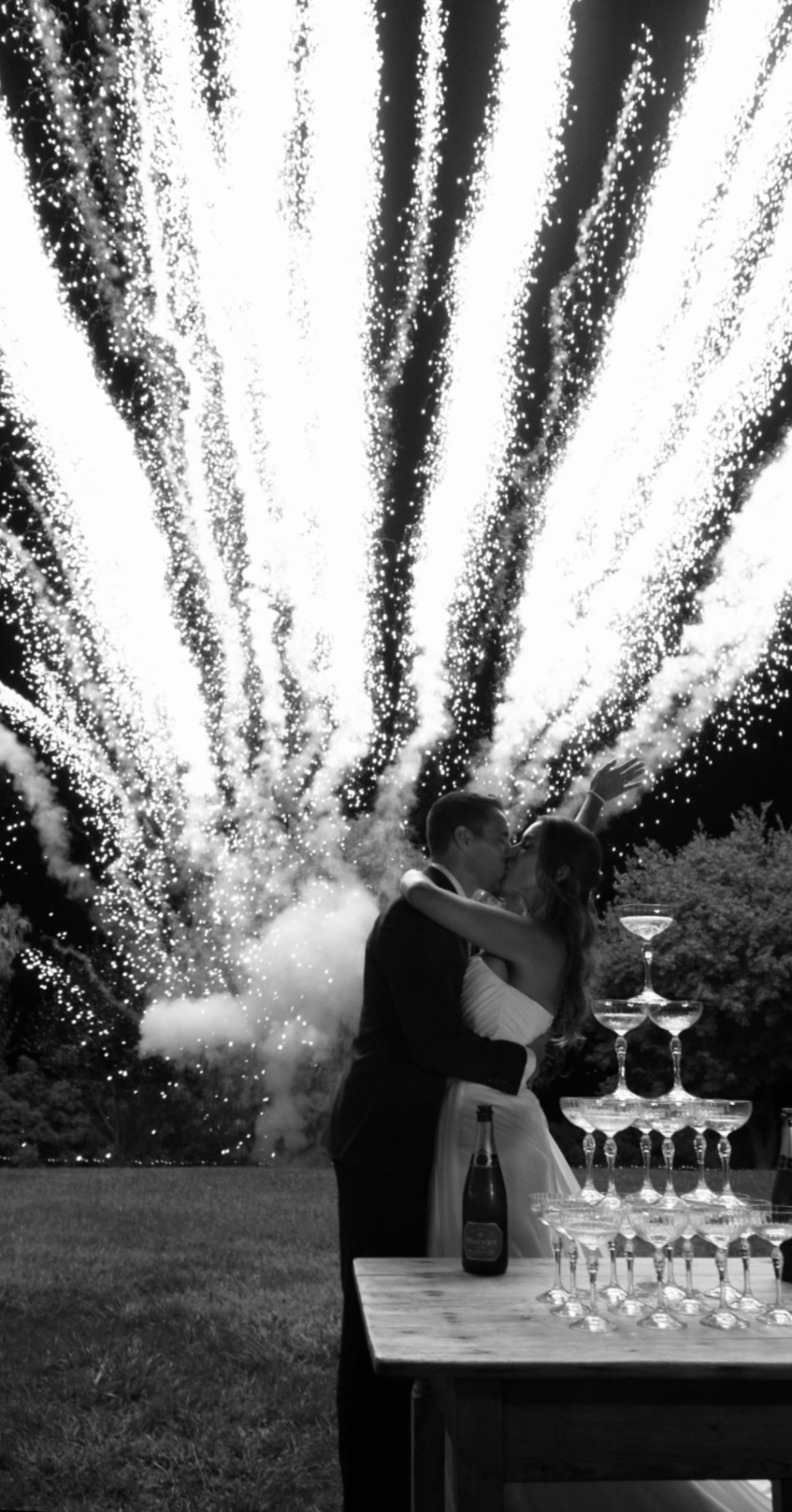 Novios besándose entre fuegos artificiales junto a una torre de copas de champagne en blanco y negro